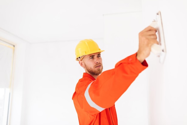 Free Photo young foreman in orange work clothes and yellow hardhat thoughtfully using putty knife at work