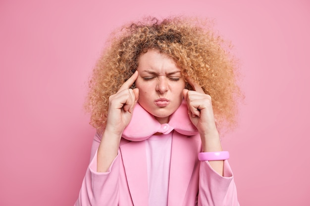 Free photo young frustrated curly haired young woman keeps fingers on temples suffers unbearable headache prepares for sleep and having rest after hard busy day