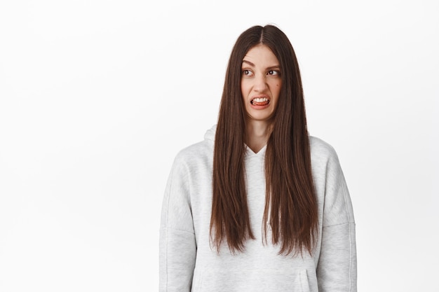 Free photo young funny girl with long hair, look disgusted aside, staring at cringe content, looking bad awful product with aversion, showing tongue and grimacing, standing over white wall