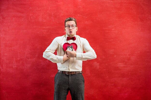 Free photo the young funny man with abstract heart and clock on red studio. concept - time to congratulate