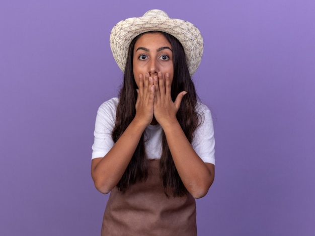 Young gardener girl in apron and summer hat  amazed and shocked covering mouth with hands standing over purple wall