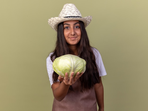 Young gardener girl in apron and summer hat holding cabbage  with smile on face standing over green wall