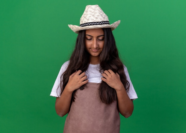 Free photo young gardener girl in apron and summer hat looking down with hands on her chest standing over green wall