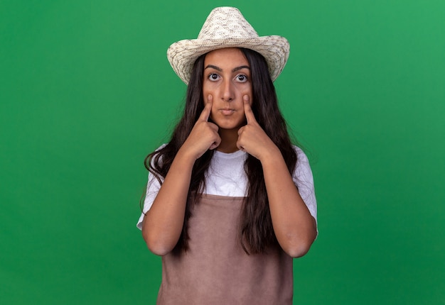 Free photo young gardener girl in apron and summer hat  surprised pointing with index fingers at her eyes standing over green wall