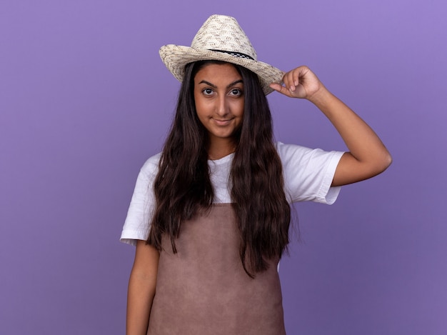 Young gardener girl in apron and summer hat  with confident smile on face standing over purple wall