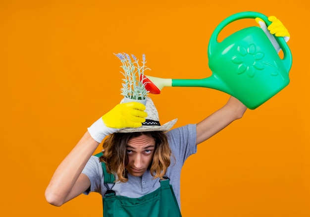 Free photo young gardener man in rubber gloves wearing jumpsuit and hat holding watering can watering plant on his head standing over orange background