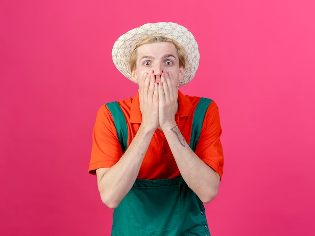 Young gardener man wearing jumpsuit and hat being shocked covering mouth with hands