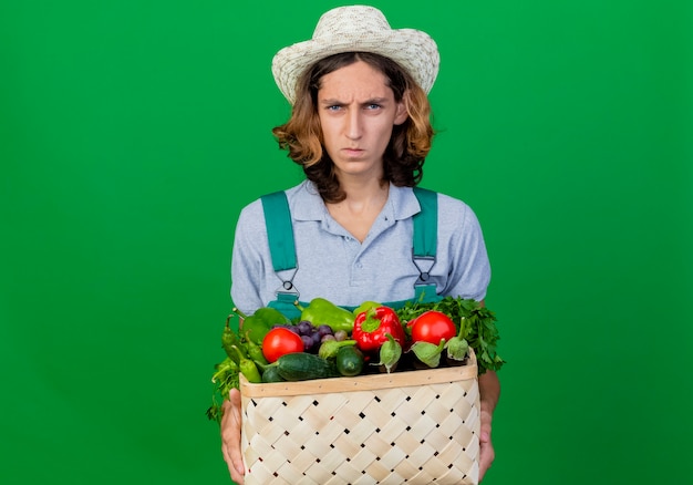 Free photo young gardener man wearing jumpsuit and hat holding crate full of fresh vegetables