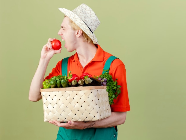 Free photo young gardener man wearing jumpsuit and hat holding crate full of vegetables biting fresh tomato standing over light background