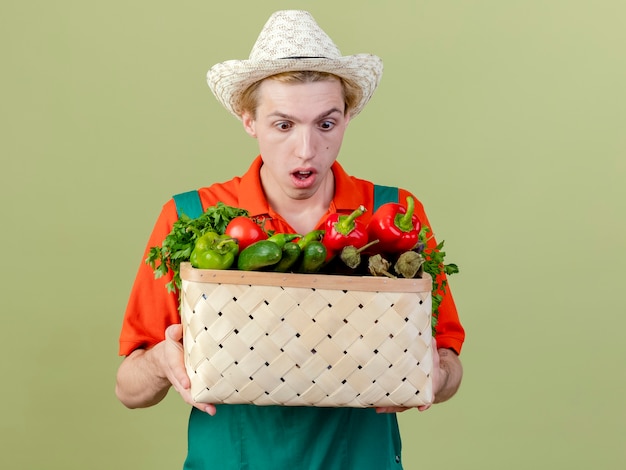 Free photo young gardener man wearing jumpsuit and hat holding crate full of vegetables looking amazed and surprised standing over light background