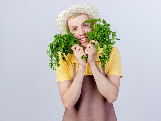 Young gardener man wearing jumpsuit and hat holding fresh herbs with smile on happy face