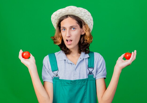 Young gardener man wearing jumpsuit and hat holding fresh tomatoes smiling