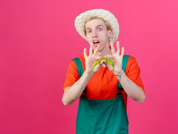 Young gardener man wearing jumpsuit and hat holding green chili pepper breaking it