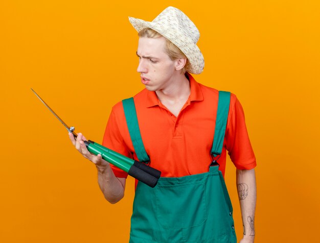 Young gardener man wearing jumpsuit and hat holding hedge clippers looking at clippers with serious face standing over orange background