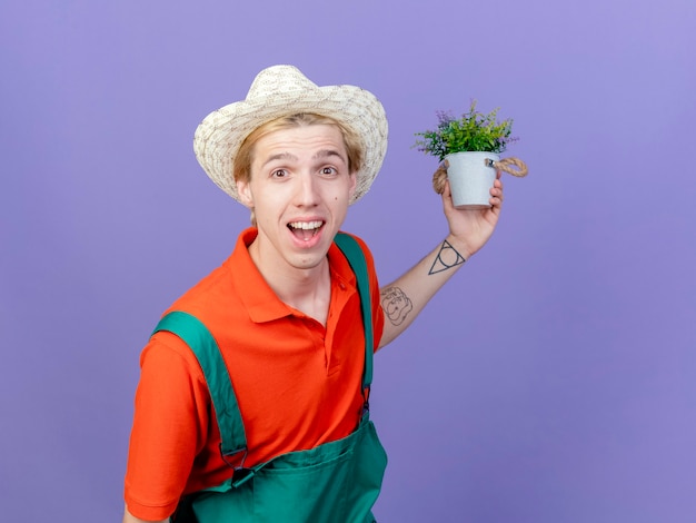 Free photo young gardener man wearing jumpsuit and hat holding potted plant looking at camera smiling with happy face standing over purple background