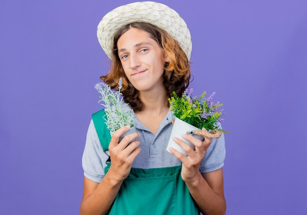 Free photo young gardener man wearing jumpsuit and hat holding potted plants