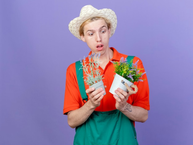 Free photo young gardener man wearing jumpsuit and hat holding potted plants