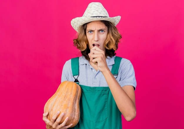 Young gardener man wearing jumpsuit and hat holding pumpkin biting cucumber