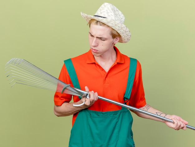 Free photo young gardener man wearing jumpsuit and hat holding rake