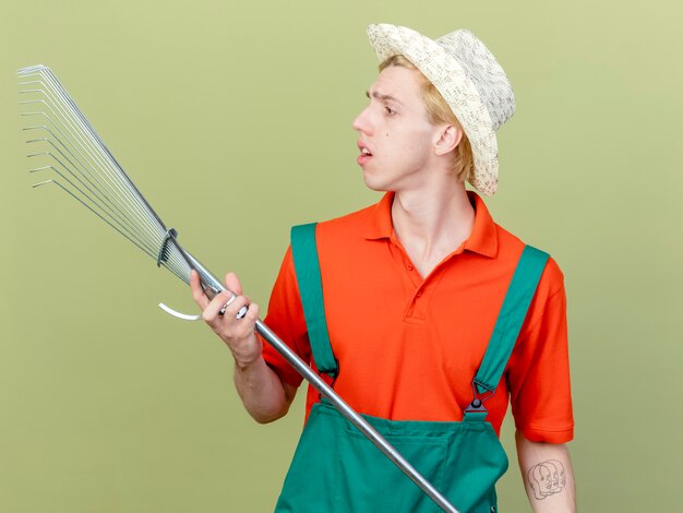 Young gardener man wearing jumpsuit and hat holding rake