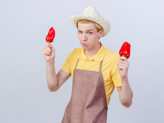 Young gardener man wearing jumpsuit and hat holding red bell peppers with skeptic smile