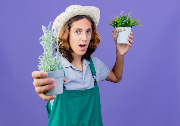 Free photo young gardener man wearing jumpsuit and hat showing potted plants smiling