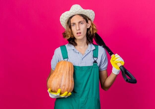 Young gardener man wearing jumpsuit and hat wearing rubber gloves holding shovel