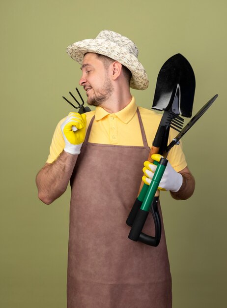 Young gardener man wearing jumpsuit and hat in working gloves holding gardening equipments looking back with smile on face 