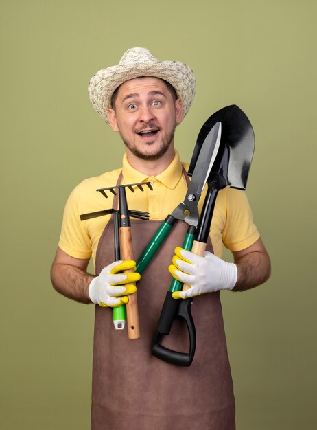 Young gardener man wearing jumpsuit and hat in working gloves holding gardening equipments looking at front smiling with happy face standing over light wall