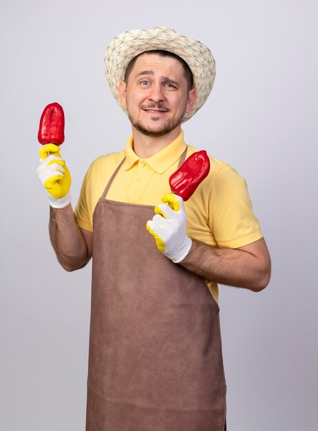 Young gardener man wearing jumpsuit and hat in working gloves holding red bell peppers, smiling with happy face standing over white wall