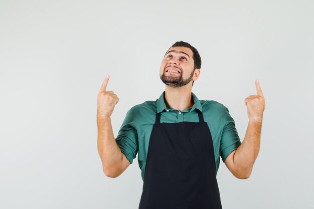 Young gardener pointing up in t-shirt,apron and looking joyful , front view.