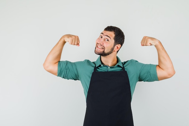 Young gardener showing his arms muscles in t-shirt,apron and looking cheerful , front view.