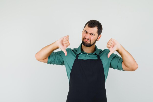 Young gardener showing thumb down in t-shirt,apron and looking displeased. front view.