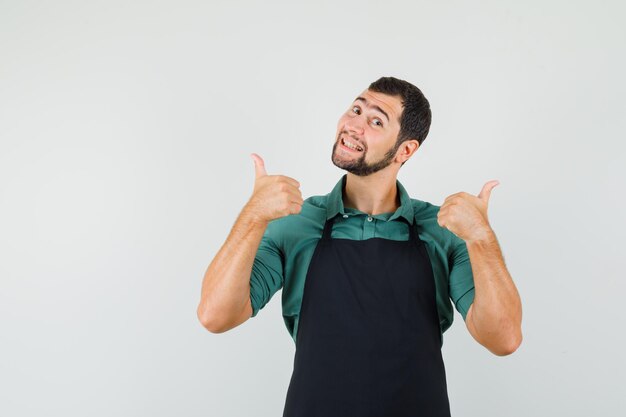 Young gardener showing thumb up in t-shirt,apron and looking happy , front view.