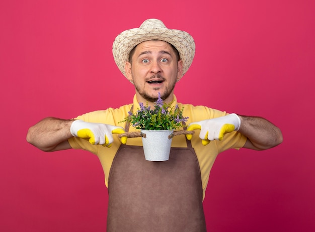 Free photo young gardener wearing jumpsuit and hat in working gloves holding potted plant looking at front with happy face smiling standing over pink wall