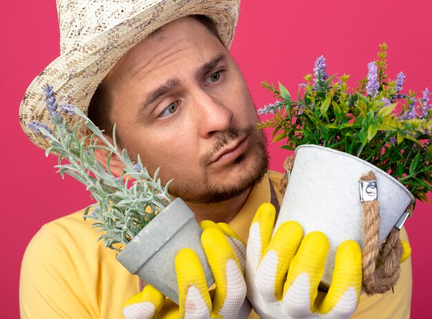 Young gardener wearing jumpsuit and hat in working gloves holding potted plants looking at them being confused standing over pink wall