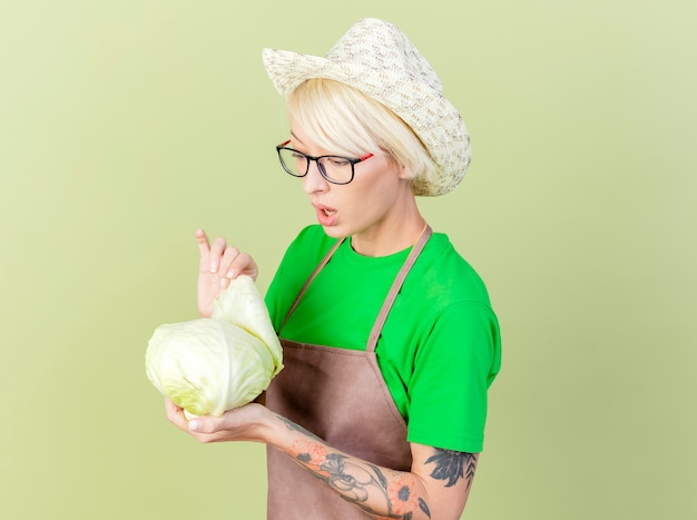 Free photo young gardener woman with short hair in apron and hat holding cabbage looking at it intrigued standing over light background