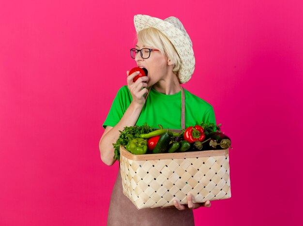 Young gardener woman with short hair in apron and hat holding crate full of vegetables