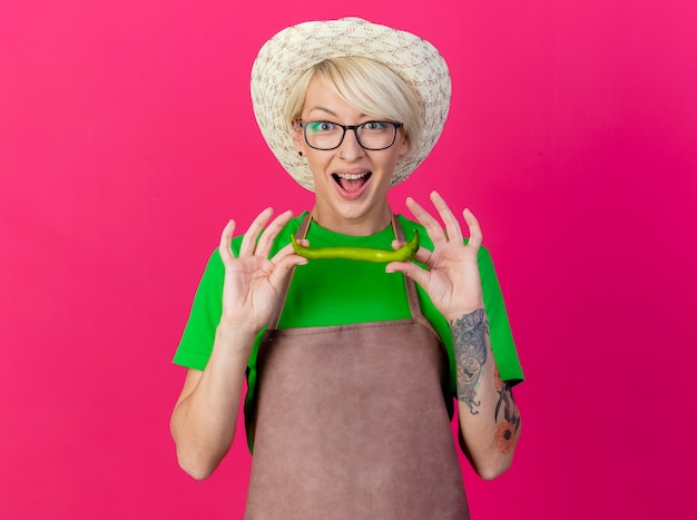 Young gardener woman with short hair in apron and hat holding green chili pepper