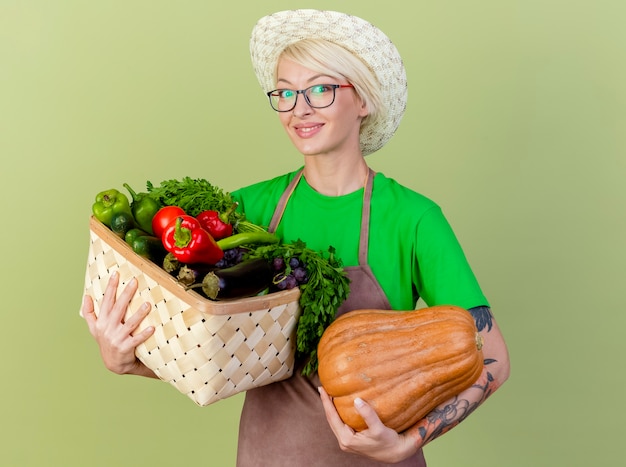 Free photo young gardener woman with short hair in apron and hat holding pumpkin and crate full of vegetables looking at camera smiling cheerfully standing over light background