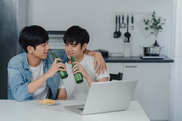 Young Gay couple drink beer while using computer laptop at modern home. Asian LGBTQ men happy relax fun using technology play social media together while sitting table in kitchen at house .