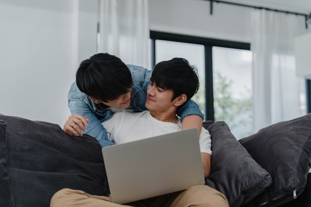 Young Gay couple using computer laptop at modern home. Asian LGBTQ+ men happy relax fun using technology watching movie in internet together while lying sofa in living room at house .