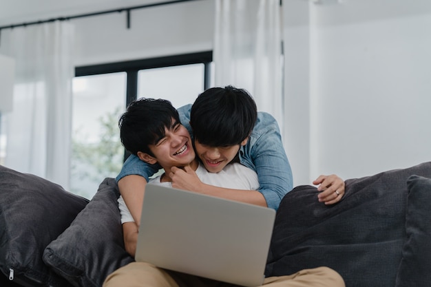 Young Gay couple using computer laptop at modern home. Asian LGBTQ+ men happy relax fun using technology watching movie in internet together while lying sofa in living room at house .