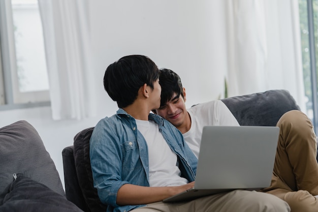 Young Gay couple using computer laptop at modern home. Asian LGBTQ+ men happy relax fun using technology watching movie in internet together while lying sofa in living room at house .