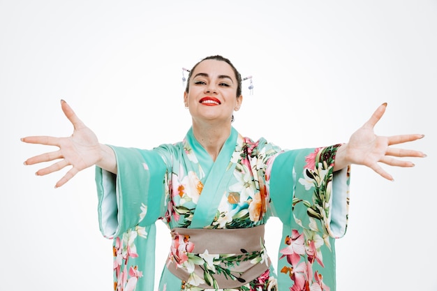 Free photo young geisha woman in traditional japanese kimono looking at front smiling cheerfully making welcoming gesture wide opening hands standing over white wall