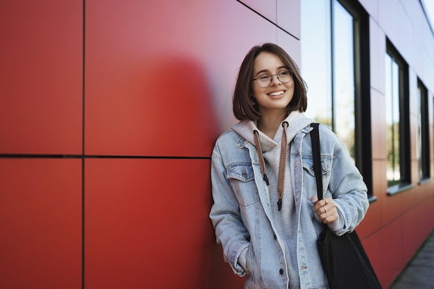 Young generation, lifestyle and education concept. Outdoor portrait of happy girl on her way home after classes, looking sideways dreamy and happy smiling, holding tote bag, lean red building.