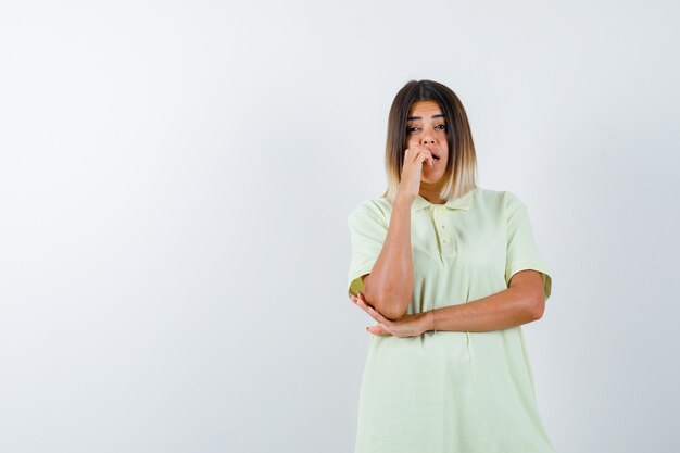 Young girl biting fingers emotionally, holding hand under elbow in t-shirt and looking scared. front view.