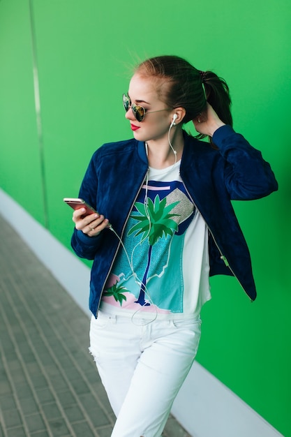 Free Photo a  young girl in a blue jacket and white pants is standing   outdoor near green wall with white line down. the girl wears sunglasses with hearts. she is leastening to music with help of headphones.