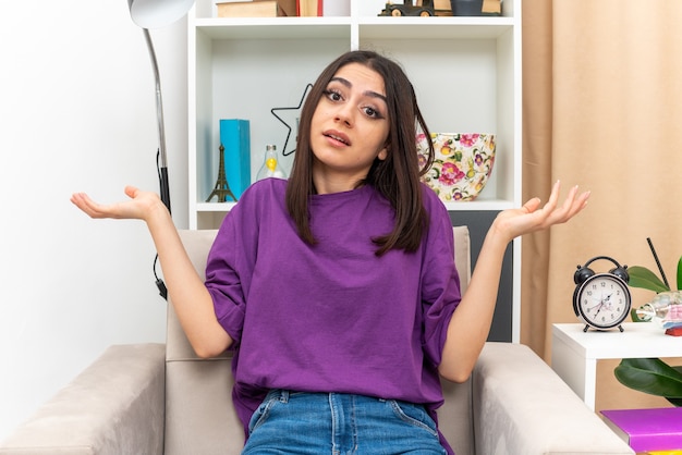 Young girl in casual clothes  confused spreading arms to the sides sitting on a chair in light living room