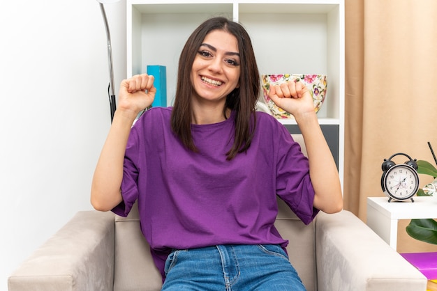 Young girl in casual clothes  happy and excited clenching fists sitting on a chair in light living room